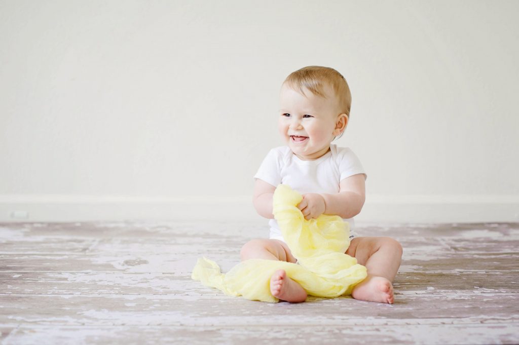 Bébé assis en body blanc qui sourit et tient une couverture jaune dans ses mains pour faire du yoga bébé